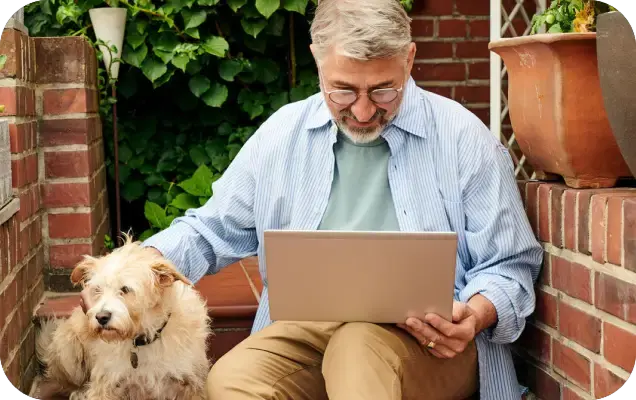 Ein älterer Mann sitzt draußen mit einem Laptop auf dem Schoß und überprüft den Bestellstatus. Neben ihm sitzt ein Hund.